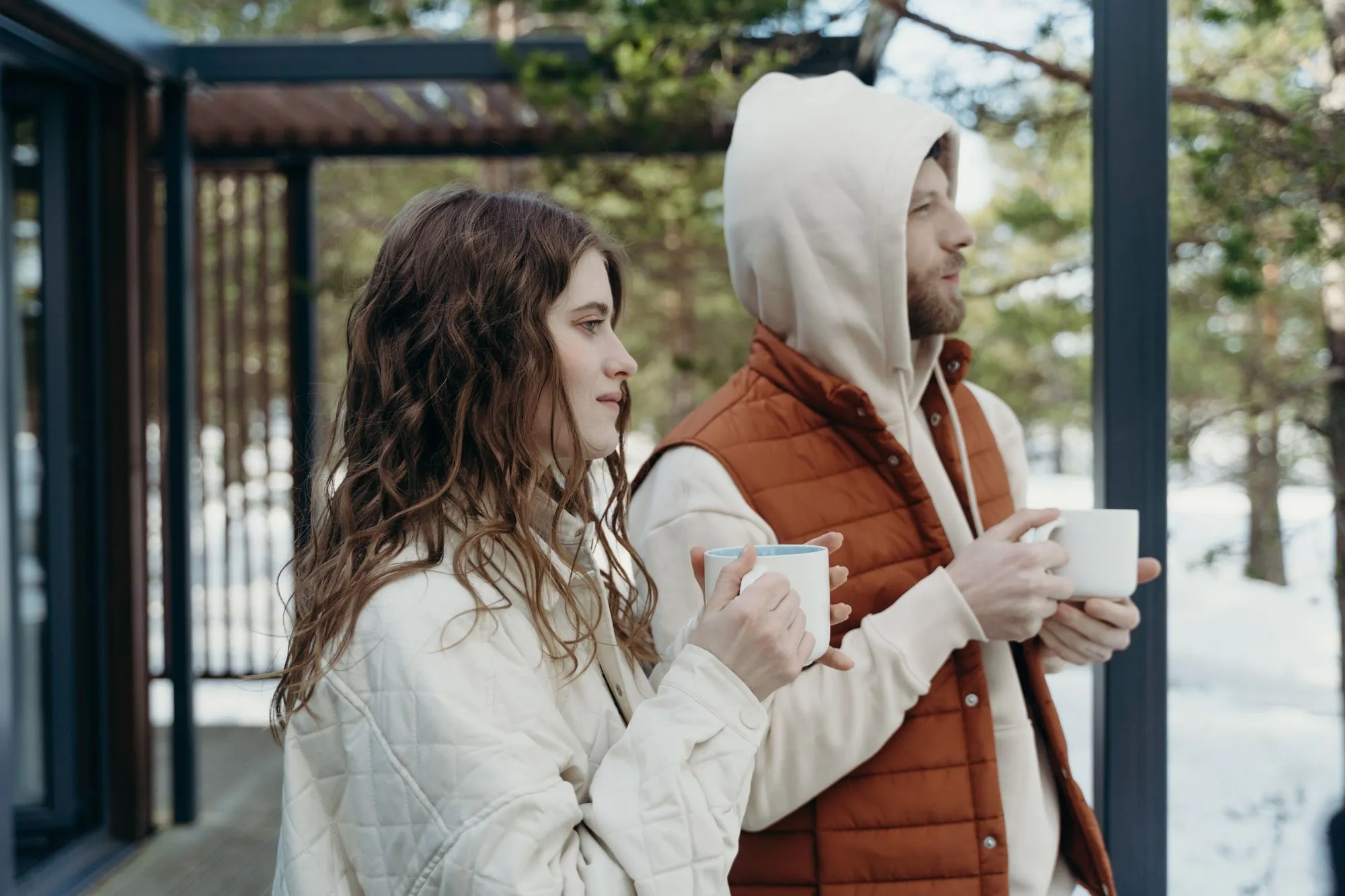 Two people holding hot drinks on a snowy porch, one in a rust quilted vest over a hoodie