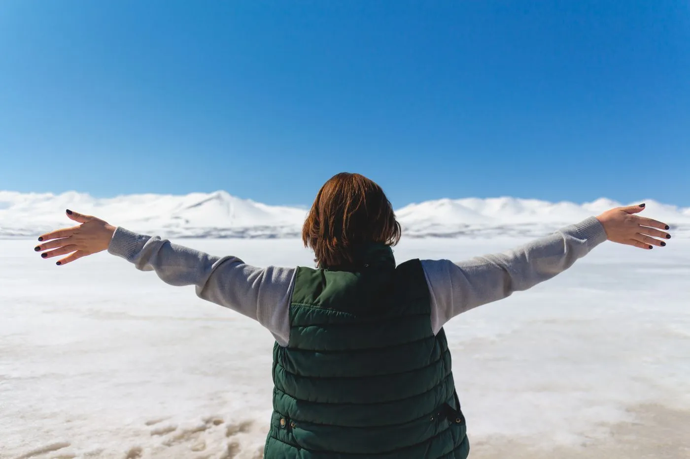 Back view of a person in a green puffer vest with arms spread wide over a snowy plain